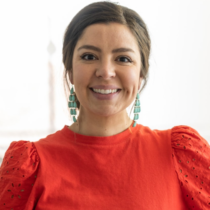 Dr. Sarah Ozuna Brown has brown hair, is wearing an orange shirt with puffed sleaves, and teal earrings. 